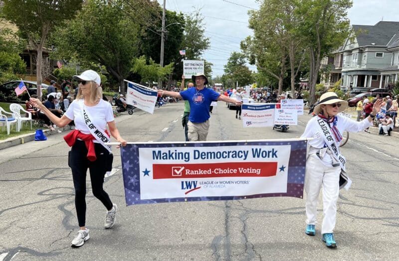 Alameda Post - two women from the League of Women Voters of Alameda walk in a parade carrying a sign. The sign says "Making Democracy Work, Ranked Choice Voting, League of Women Voters of Alameda"