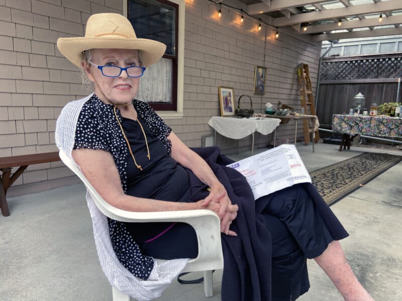 Alameda Post - a woman in a straw hat and blue glasses reclines in a white lawn chair and smiles at the camera. The woman is Gretchen Lipow.