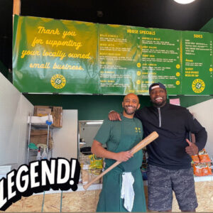 Alameda Post - two men stand together at a hotdog shop. One is holding a signed baseball bat that he appears to have been given by the other