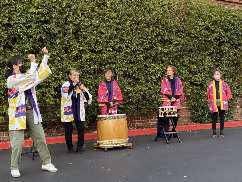 Alameda Post - five people in colorful garments stand in front of a brick wall covered in leaves. Two of the people have percussion instruments. 