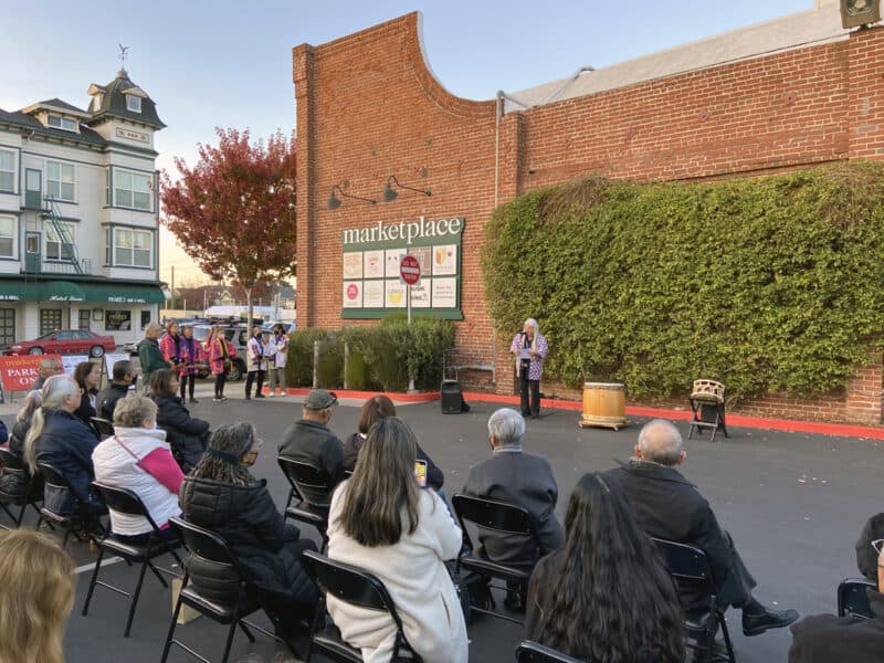 Alameda Post - a crowd is seating watching a presentation outdoors in a parking lot. The speaker stands in front of the brick side of the Alameda Marketplace