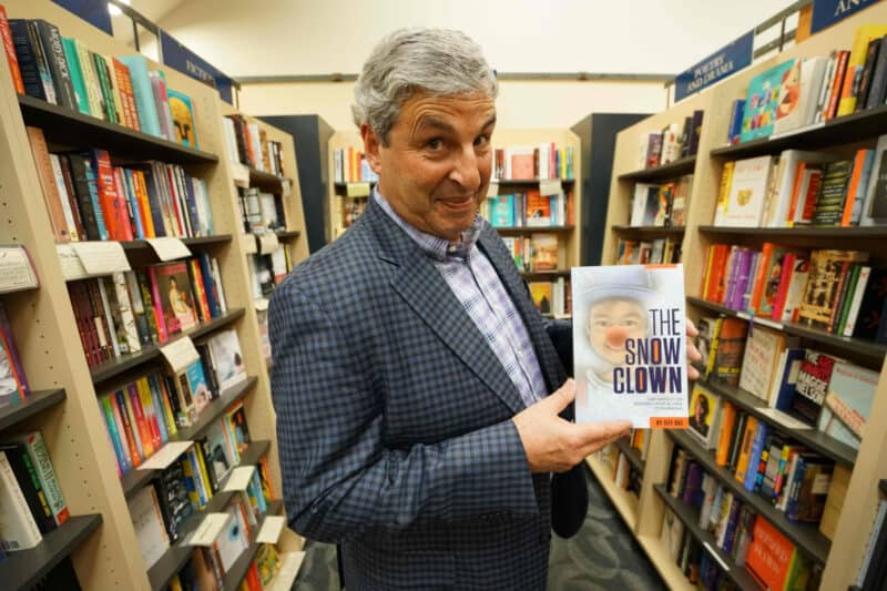 Alameda Post - a man in a blue suit stands among racks of books. He holds one of them and smiles at the camera, displaying the book