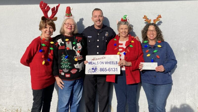 Alameda post - four people smile and stand together holding a sign saying "Alameda Meals on Wheels. 865-6131"