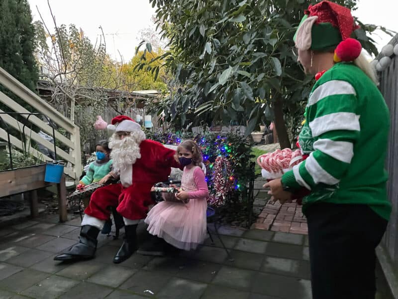Alameda Post - Santa visits a family and sits in a backyard patio and poses for a picture with two children, one in a frilly pink dress and the other in a striped sweater. An elf looks on.