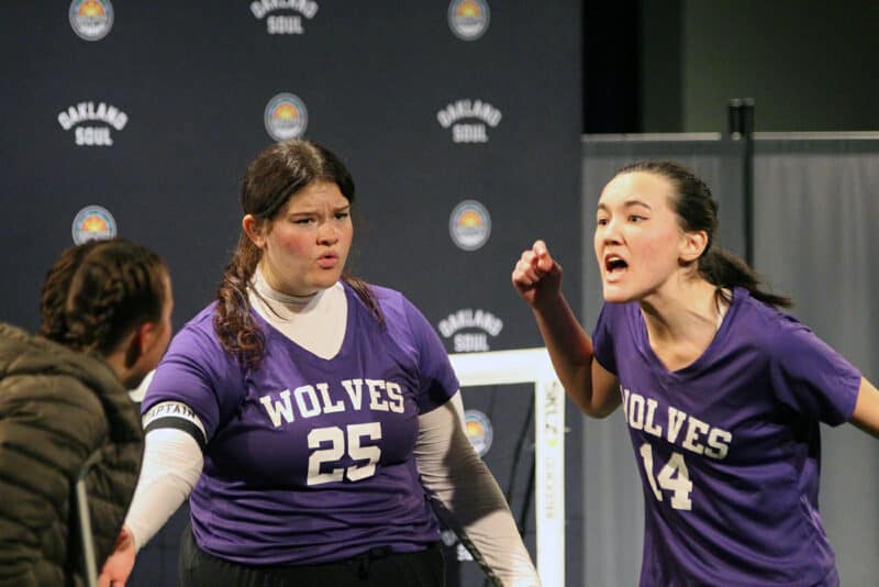 Alameda Post - three actors stand onstage in this close up photo. Two are wearing purple soccer uniforms, and one is wearing a puffer jacket. One of the soccer players is yelling at the person in a jacket. The two jerseys are for The Wolves team