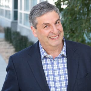 Alameda Post - a man in a blue suit and checkered blue shirt smiles at the camera. This is Jeff Raz's headshot.