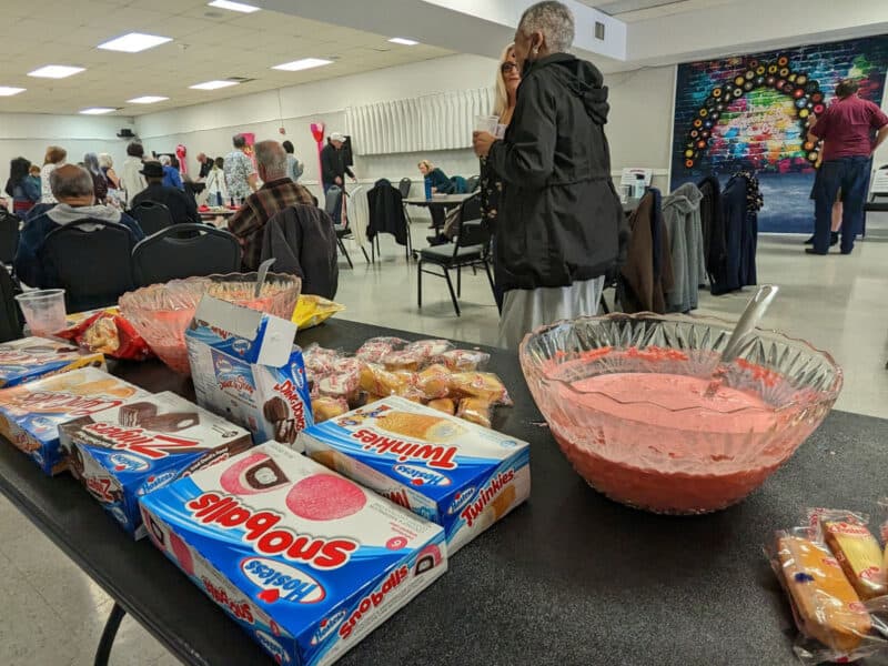 Alameda Post - the refreshments table is laid with big bowls of red bunch and hostess snacks including snowballs, twinkies, Zingers, ding dong, and orange cream cupcakes. Bags of chips peak out from behind a second punch bowl. In the background, the Self Station is set up, and people sit at tables or dance on the dance floor. 