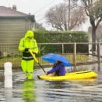 Alameda Post- A person in rain gear pulls a child holding an umbrella in a kayak along flooded streets