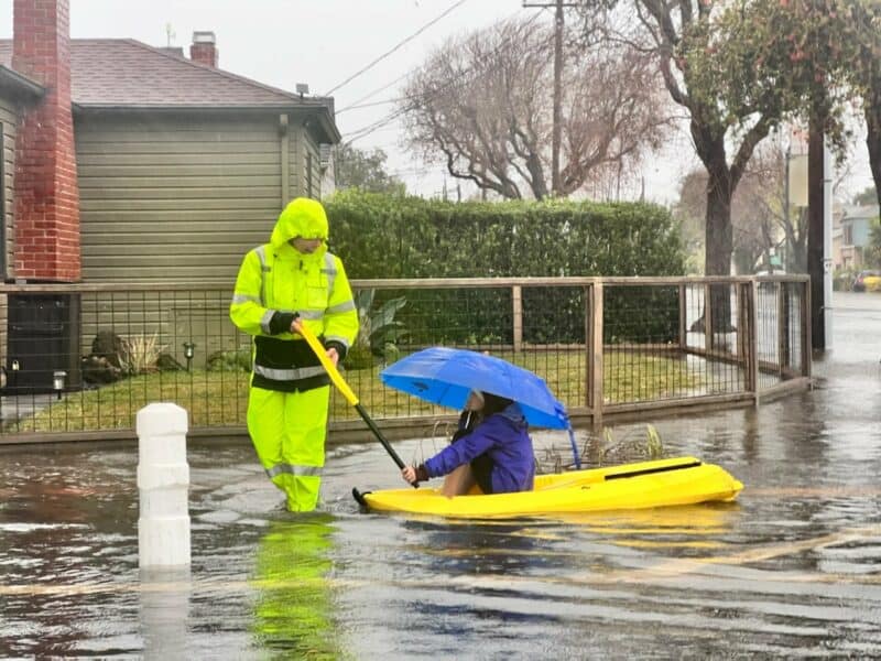 Alameda Post- A person in rain gear pulls a child holding an umbrella in a kayak along flooded streets