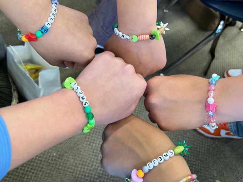 Alameda Post - a close up photo of the wrists of five high school students. They are wearing beaded bracelets that say "Swell"