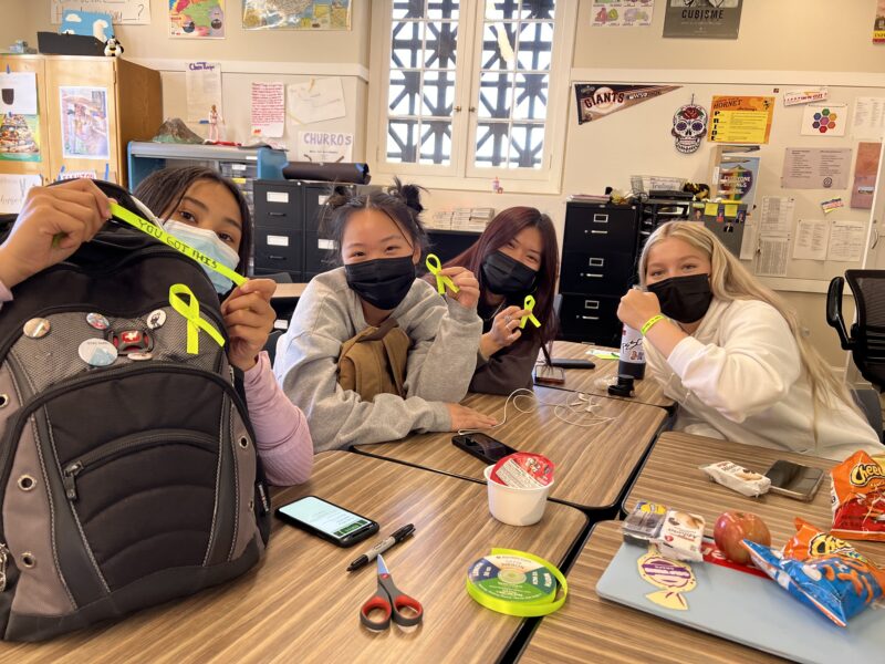 Alameda Post - high school students sitting at their desks look at the camera and show off bracelets saying "You got this" and bright yellow ribbons