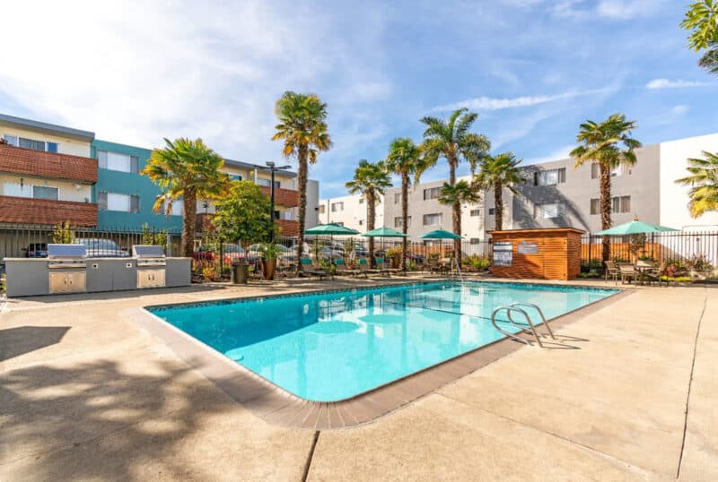 Alameda Post - a pool at the Rica Vista apartments. The apartment buildings, patio furniture, umbrellas, and palm trees frame the background.