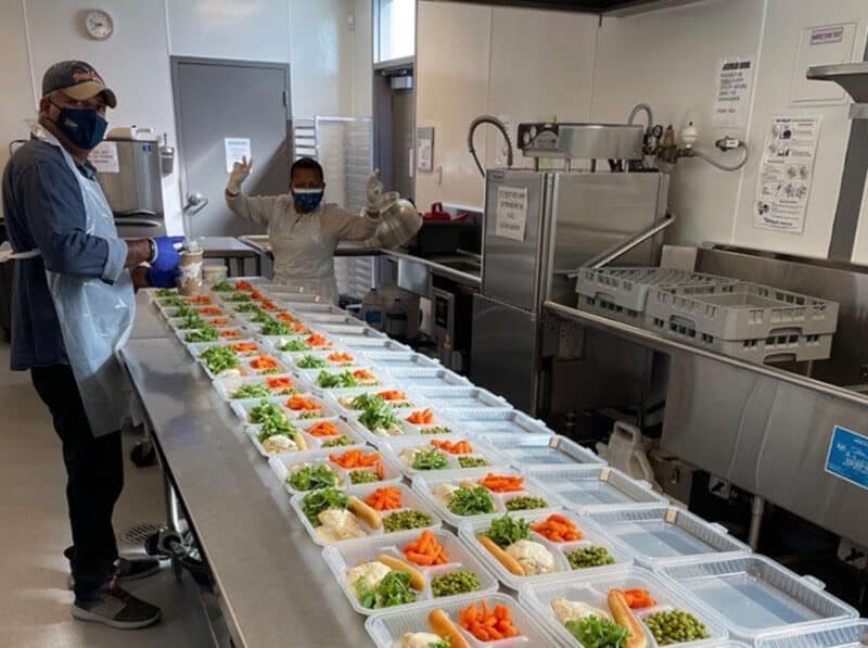 Alameda Post - workers in a kitchen package up loads of food into individual meals. The containers are reusable and part of the StopWaste Reusable Foodware Grant materials