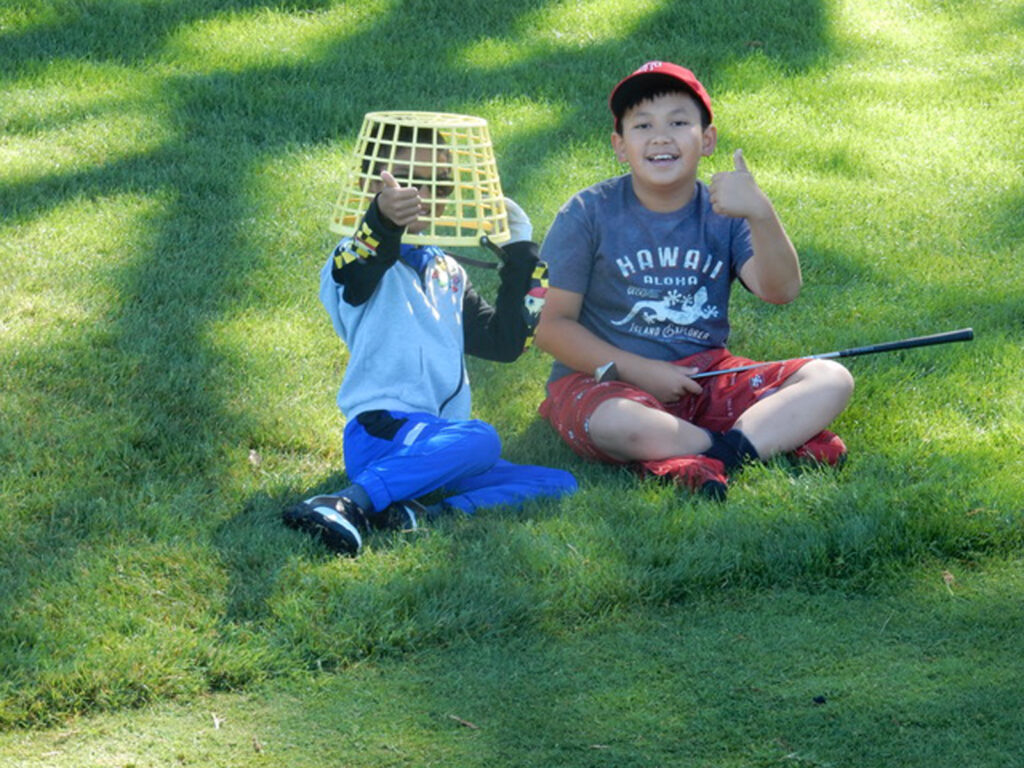 Alameda Post - two young golfers from the Alameda Junior Golf Association sit on a green lawn. One holds a golfclub and the other has a basket over their head