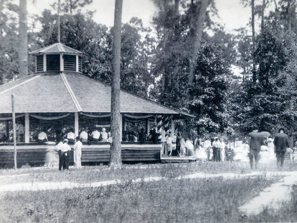 Alameda Post - an old black and white photo of an outdoor wooden, open air pavilion. People in suits and dresses are in and around it.