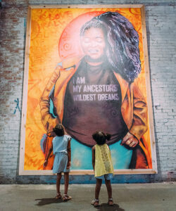 Alameda Post - two children look at a mural of a woman wearing a shirt that says "I am my ancestors' wildest dreams"