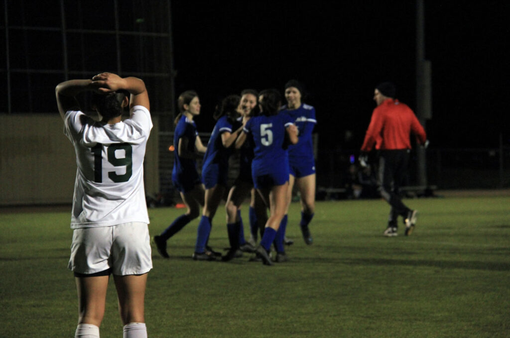 Alameda Post - a group of Encinal Jets soccer players in blue jerseys celebrate in a cluster while a member of the opposite team looks on with their hands behind their head.