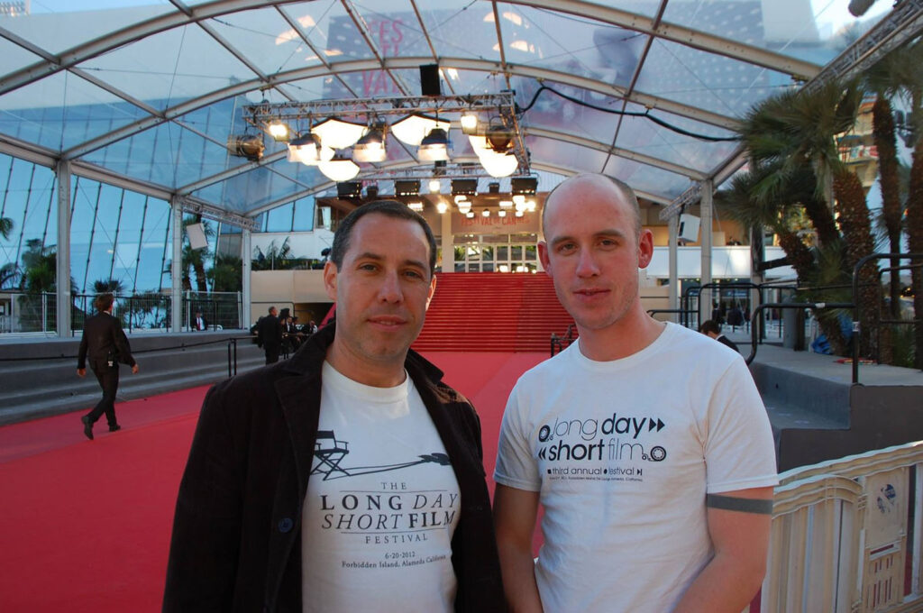 Alameda Post - Mark Farrell and Colin Blake pose in a large indoor room by a red carpet wearing THe Long Day Short Film Festival T shirts