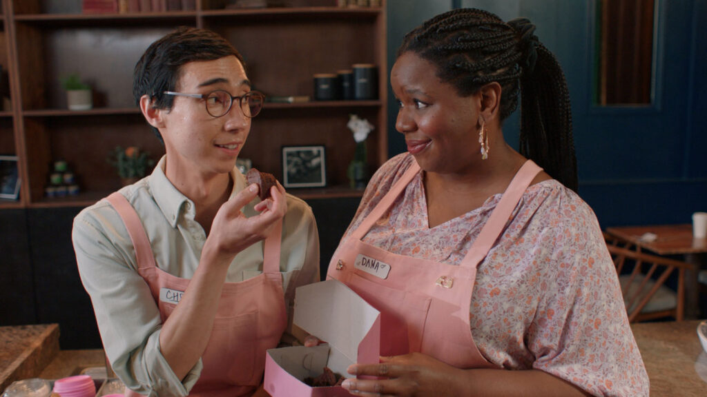 Alameda Post - two actors stand next to each other and smile slightly at each other over a box of treats