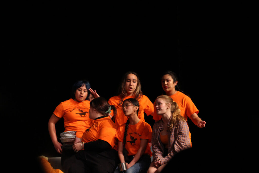 Alameda Post - a group of actors in bright orange shirts against a black background in The Lightning Thief