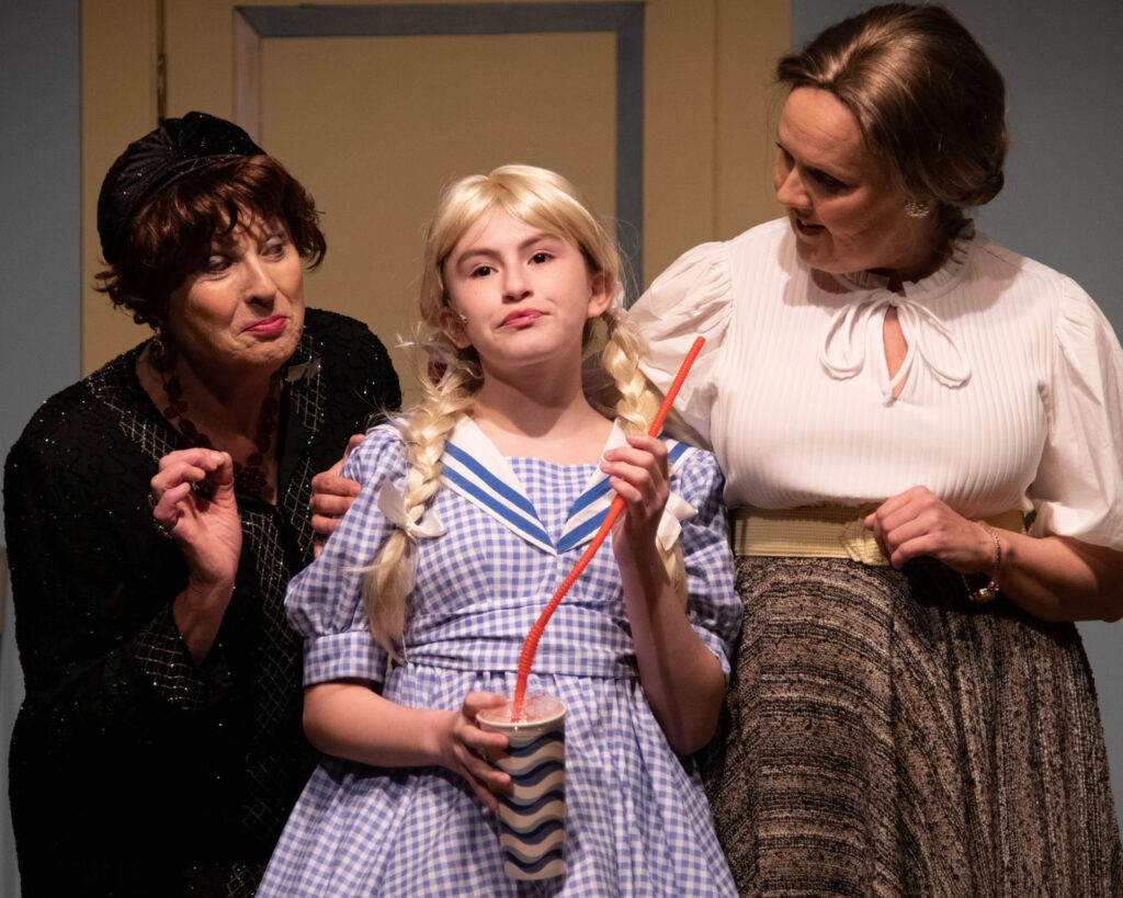 Alameda Post - three actors in a row onstage in Ruthless! The Musical. In the center, an actor in a blue gingham dress holds a soda with a very long straw