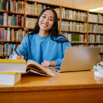 Alameda Post - a student sits at a desk in the library and smiles. Read on for summer internship details.