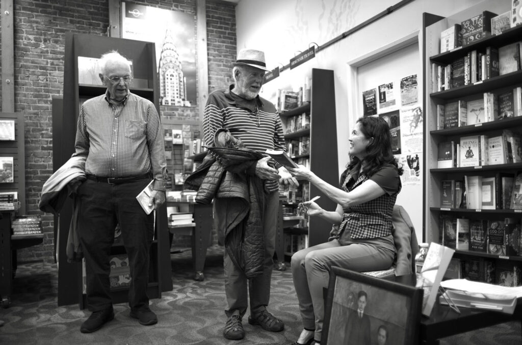 Alameda Post - two men stand and talk and shake hands with Karin Jensen at her book signing 