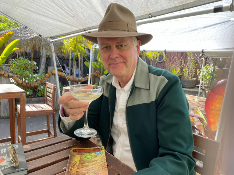 Alameda Post - Eddie Muller sits outside at a table at Forbidden Island and holds up a cocktail