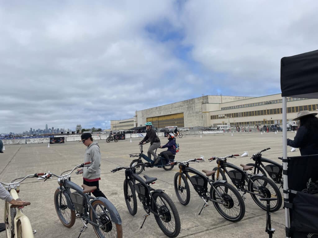 Alameda Post - two people test out E-bikes in a large concrete area. A row of empty E-bikes waits for testers