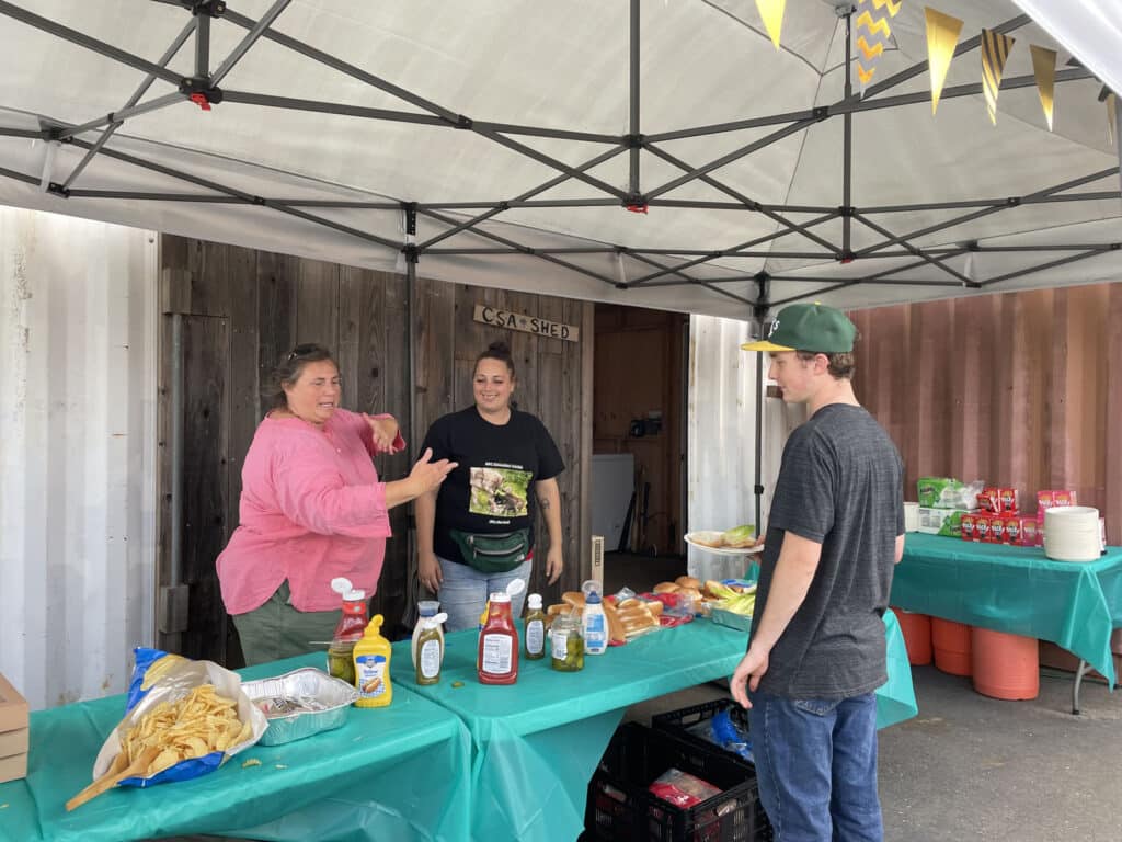 Alameda Post - Three people stand around a table with chips, condiments, and buns at the Farm2Market Harvest Festival