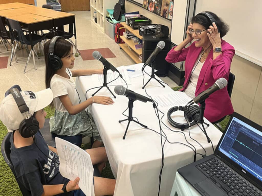 Alameda Post - two 4th grade podcasters from Little Minds Big Talks sit at a table in their classroom. The mayor sits across from them and smiles