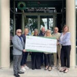 Alameda Post - seven people stand in front of an AMP building holding a large check for Power Up for Learning