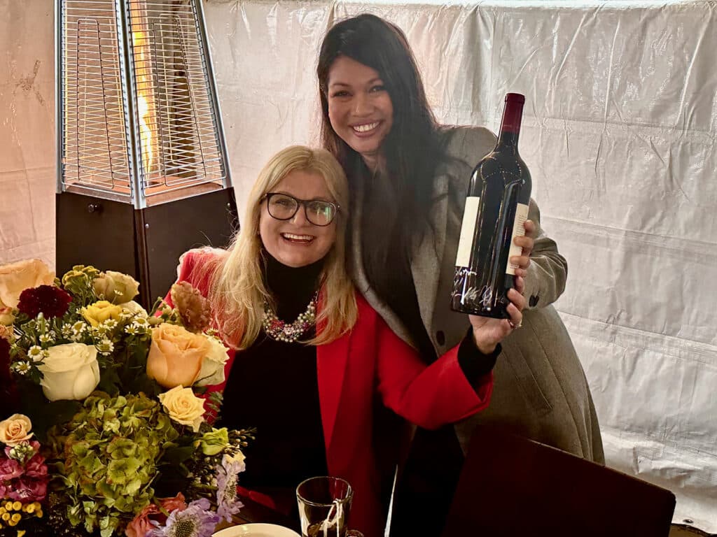Alameda Post - at a restaurant, Kelly Lux holds up a bottle of wine and smiles while Joann Guitarte poses next to her