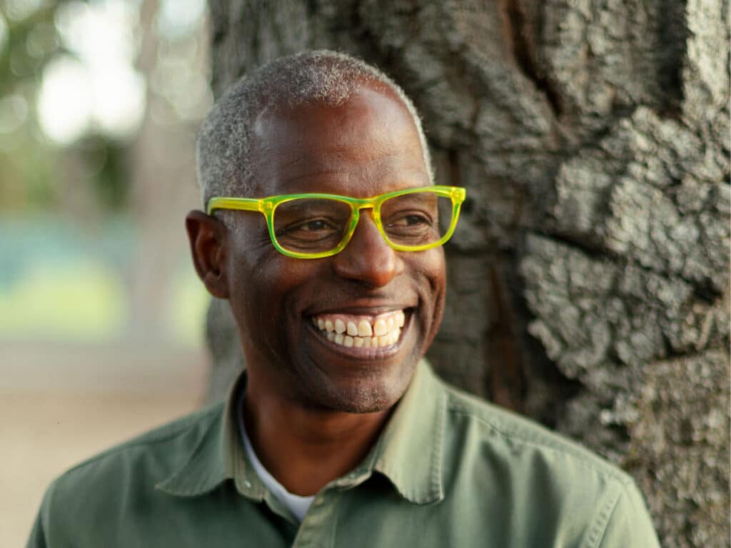 Alameda Post - Amos White smiles in front of a tree wearing bright yellow rimmed glasses