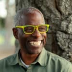 Alameda Post - Amos White smiles in front of a tree wearing bright yellow rimmed glasses