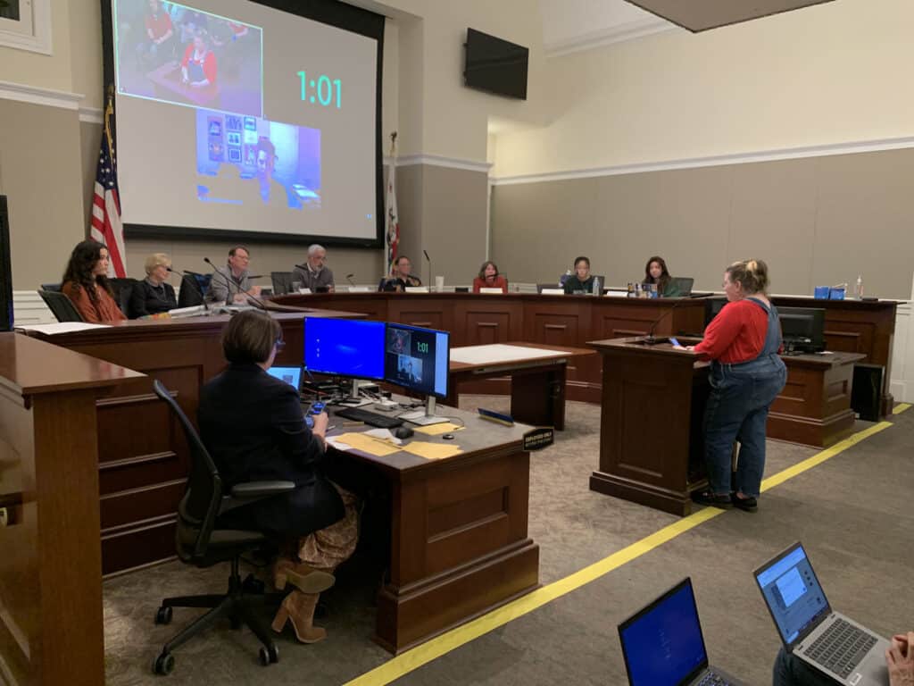 Alameda Post - a woman with a red shirt stands before the AUSD school board and talks