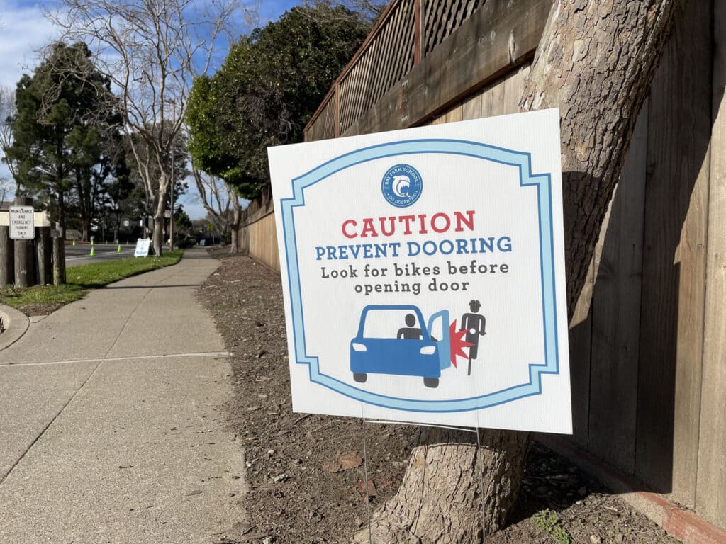 Alameda Post - a sign that says "Caution. Prevent dooring. Look for bikes before opening doors." A little graphic of a car and cyclist shows them getting doored.