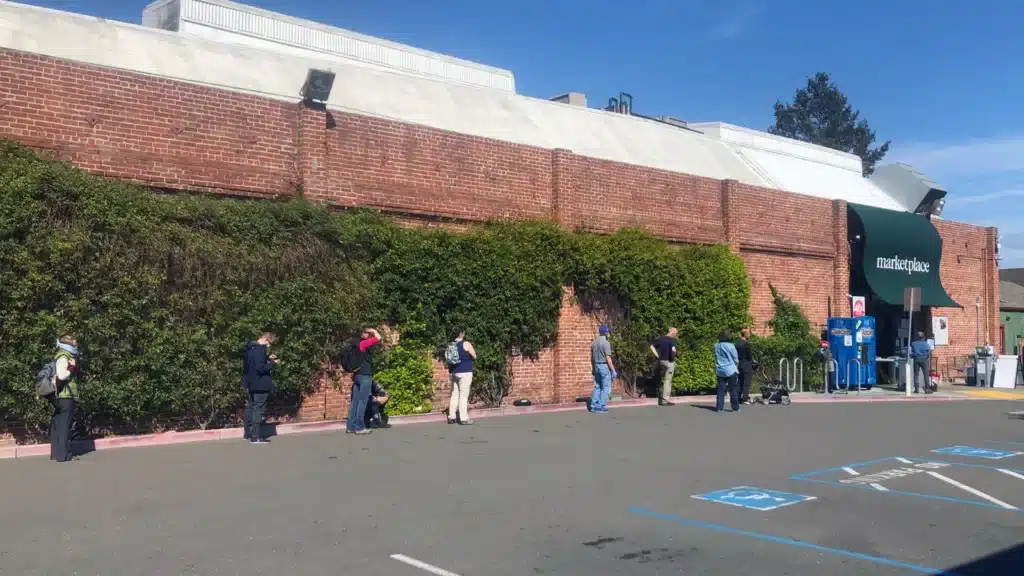 Alameda Post - A widely-spaced line of people wait to enter the Alameda Marketplace during the COVID-19 pandemic