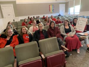 Alameda Post - AUSD teachers wearing red sit in the audience of a board meeting. One teacher holds a sign that says, "I support my students. I should be able to support my family."