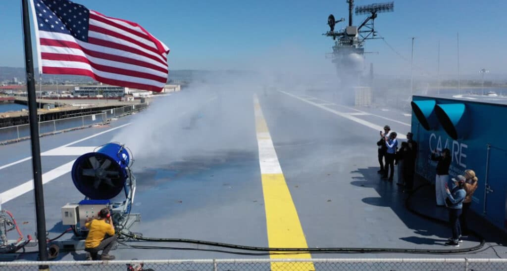 Alameda Post - on the deck of the USS Hornet, an American flag waves while people use a very large fan apparatus to spray salt and water across the deck