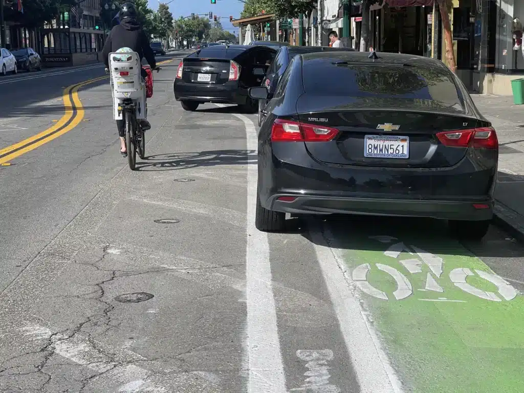 Alameda Post - a woman riding a bike with a child on the back swerving outside the bike lane because cars are parked in the lane, obstructing it.