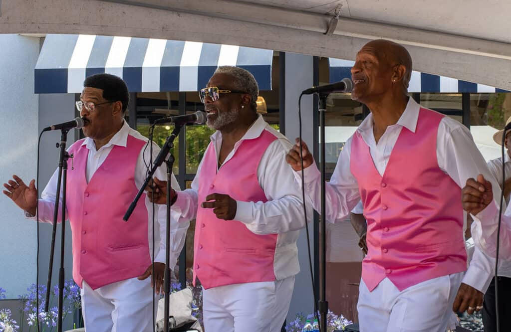 Alameda Post - three members of The Best Intentions wear matching bright pink vests and white dress shirts. They stand up to microphones and sing and snap their fingers