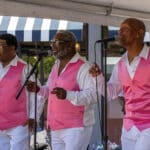 Alameda Post - three members of The Best Intentions wear matching bright pink vests and white dress shirts. They stand up to microphones and sing and snap their fingers