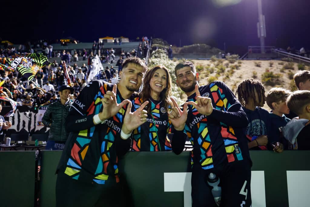 Alameda Post - two Oakland Roots players flash a "W" and stand with a woman in the stands