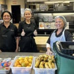Alameda Post - three women stand in an AUSD kitchen and smile. They are wearing gloves and bonnets