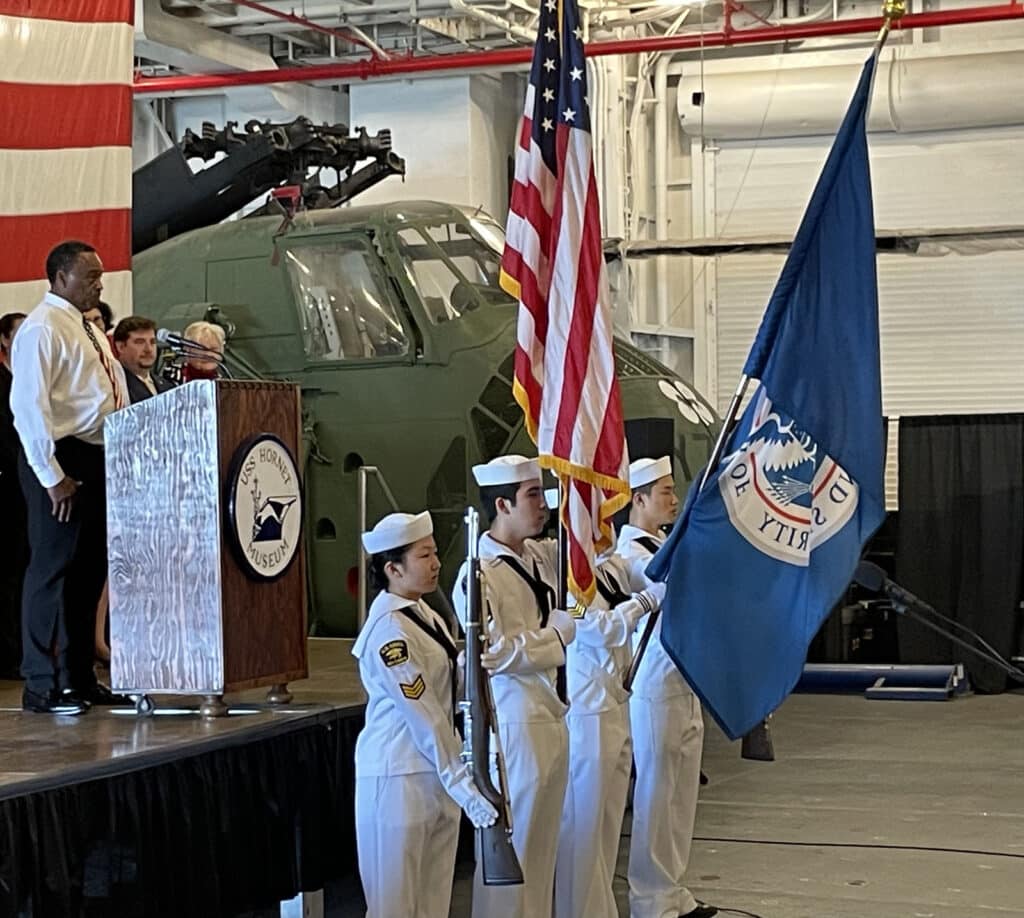 Alameda Post - aboard the USS Hornet, people in uniform present the flags while someone on stage sings the national anthem