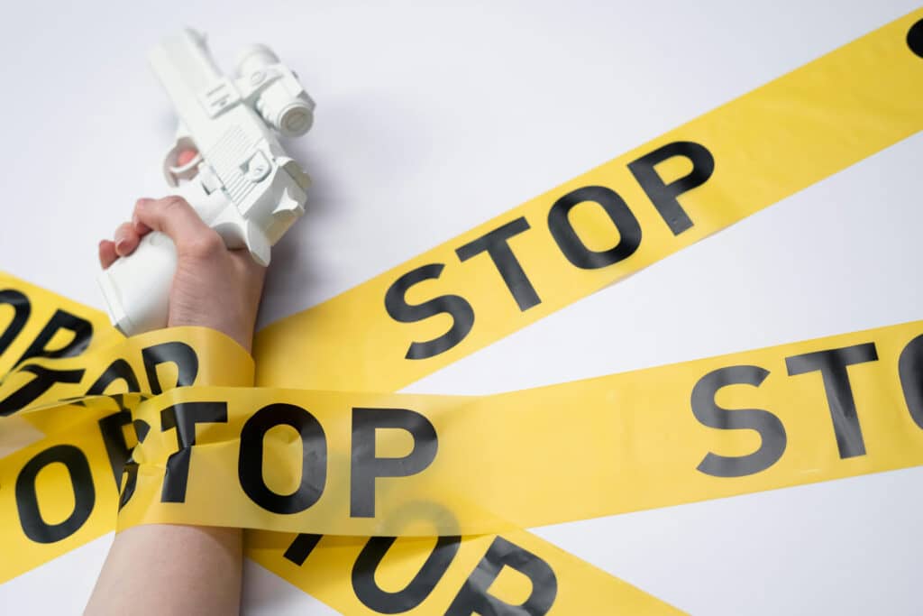 Alameda Post - a photo of a hand holding a prop gun. The hand is held down to a surface by long strips of yellow and black tape that says "Stop."