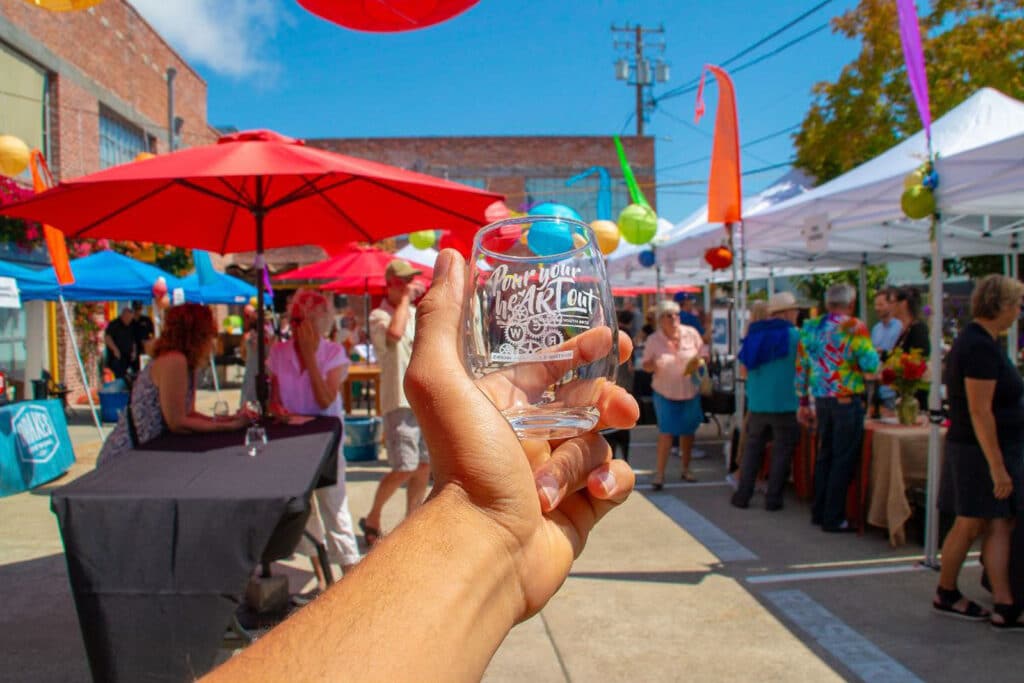 Alameda Post - a hand holds out a wine glass that says "Pour Your Heart Out." In the background, people mill around a colorful festival