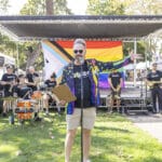 Alameda Post - a man in bright rainbow accessories stands in front of the Alameda Pride stage and introduces a band