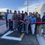 Alameda Post - a group of people smile for a photo as Marilyn Ezzy Ashcraft holds a large pair of scissors to cut the ribbon on a new section of the Cross Alameda Trail